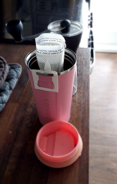 A large pink travel coffee cup sits on a wooden benchtop with a pourover bag fitted, partially filled with ground coffee. Behind it is a Hario Slim coffee grinder.