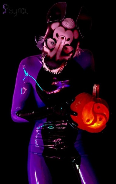 Purple latex pup Styra stands only wearing their purple latex catsuit, without restraints or collars.  They wear a skeleton pup hood made by Grunge Bunny.  With a menacing look, they hold a pumpkin carved with a triskelion.