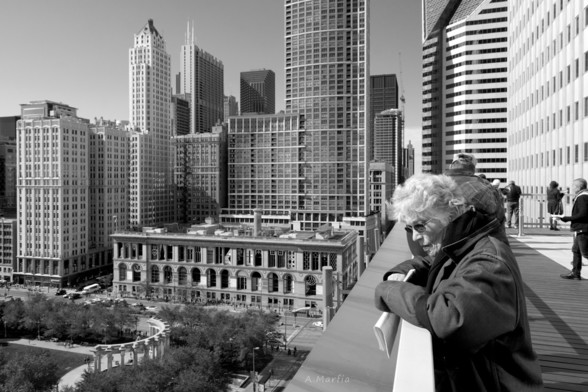 A black and white photograph taken from the 11th floor balcony of the Prudential Building during Open House Chicago 2015. Pictured is a women looking over the edge with part of Millennium Park, the Chicago Cultural Center, and other buildings along Michigan Avenue in the background.
