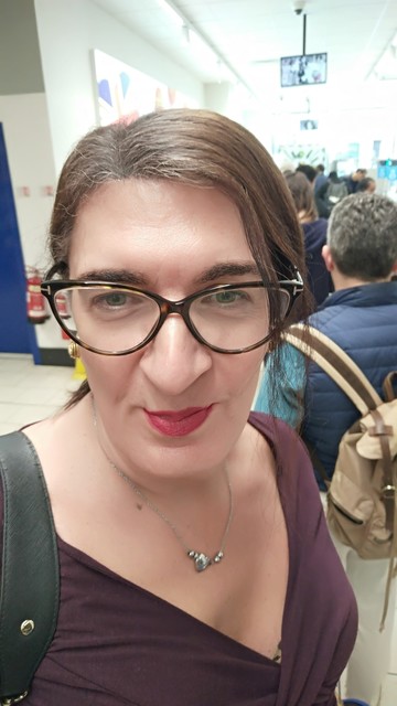 A selfie of a brunette trans woman in a wine colored top wearing ING glasses with a queue in the background