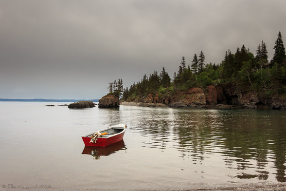 A scene of a shore with rocky cliffs and evergreens and bushes etc on the top. There are some rocks sticking up from the surface of the water, one fairly high with steep sides and skinny evergreens growing from the top of it. In the foreground is a red small motorboat, with the motor resting up on the stern. It is in the middle of the water. The sky is a flat grey with darker lower areas.
