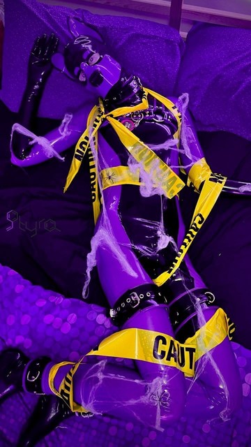 Pup Styra laid on a bed, limbs sprawled.  Theyâ€™re covered in yellow CAUTION tape and spiderwebs. Theyâ€™re wearing tight, figure fitting purple and black latex.