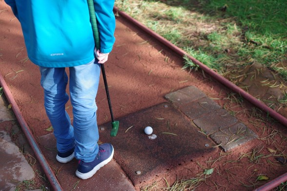Photo of legs and feet of kid holding mini golf club, standing next to the mini golf tee.