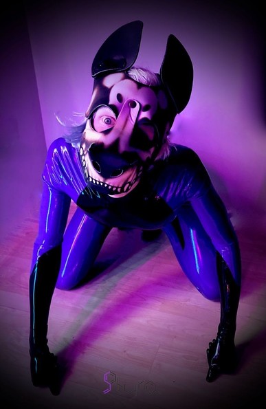 Latex Pup Styra is kneeling on a floor, both front hands out in front sat dog-like.  They are wearing their purple and black latex and wearing a skeleton puppy hood.
