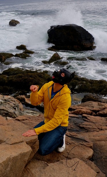 A hooded human dog in yellow hoodie has his tongue out while kneeling on the brown rocks over the foamy Atlantic Ocean