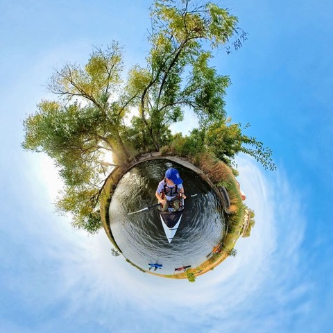 Photo of a person kayaking on a lake, watching as other kayakers enter the lake. In the background are green & yellow trees under a blue sky with wispy white clouds.