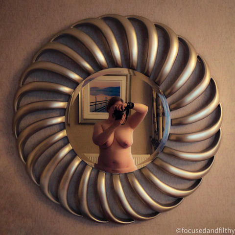 Square colour photograph of a large round ornate mirror. The edges have curved lines like a shell all the way round. In the mirror I’m stood naked holding my large digital camera up to my face to take the image. You can just see my torso.