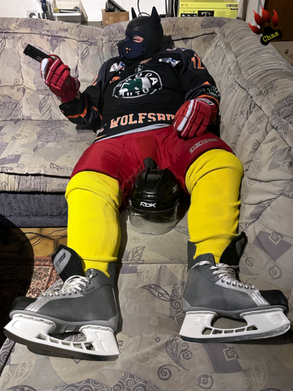 human pup, geared up in ice hockey gear, lying on sofa, remote control in hand