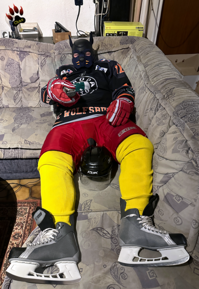 human pup, geared up in ice hockey gear, lying on sofa, remote control aimed at viewer in hand
