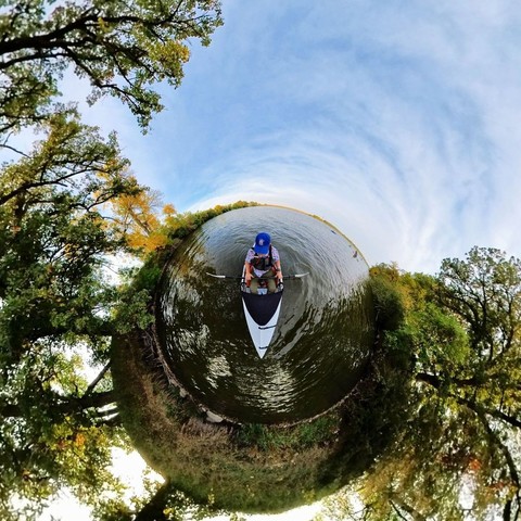 Photo of a person kayaking on a lake, pausing along the shoreline to view the trees beginning to turn autumn colors.