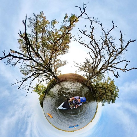 Photo of three people kayaking on a lake, past two mostly leafless trees along the shoreline. Above is a light blue sky filled with wispy white clouds.