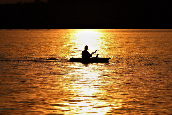 A kayaker in silhouette on a lake surface lit by sunset light with a white streak down the center and golden light reflecting off the ripples.