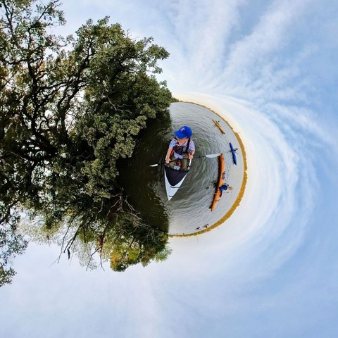 Photo of several kayakers on a lake, paddling alongside trees on the shoreline. The light blue sky is filled with wispy white clouds.