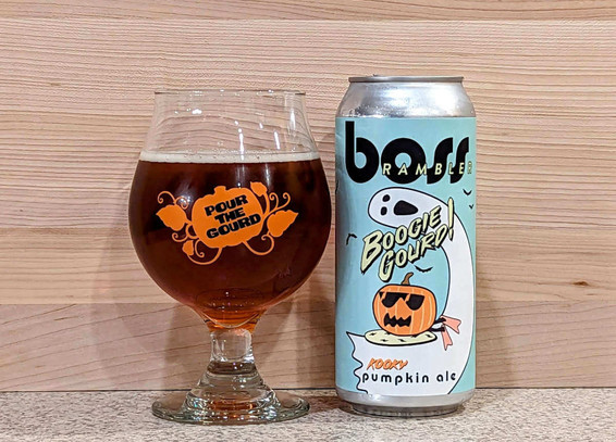 A snifter glass of Boogie Gourd kooky pumpkin ale from Boss Rambler Beer Club of Bend, Oregon. The glass has a logo of an orange pumpkin and leaves with the phrase "Pour the Gourd" in the center, while the can has a cartoonish jack-o-lantern that looks like it's wearing sunglasses paddling on a boogie board but the wave it's surfing is actually a ghost.