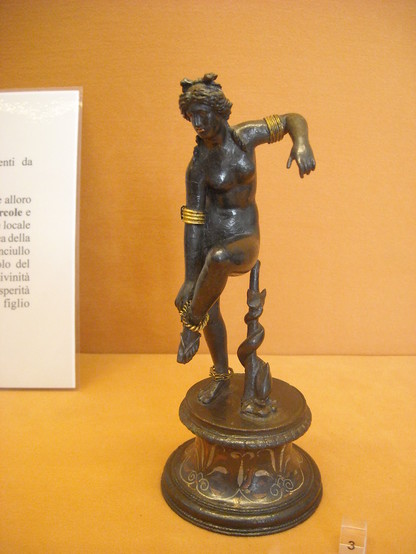 Statuette of Venus or Aphrodite from Pompeii. She is depicted in a dynamic pose, balancing on one foot as she puts on a golden anklet. Her left arm is raised and angled back for balance. The goddess is nude with the exception of her jewellery, two golden anklets and several arm rings on each side. She also wears sandals. The bronze is dark, making the golden jewellery stand out in contrast. Her lips, probably made of copper, shine in a seductive red.