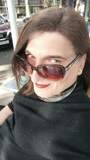 A brunette trans woman dressed in black with a wire and gold choker necklace peers above her sunglasses