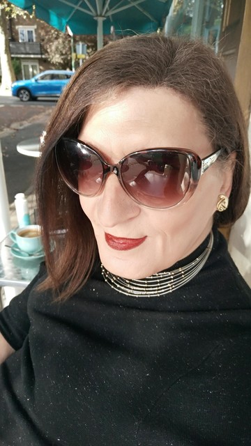 A brunette trans woman dressed in black with a wire and gold choker necklace wearing brown sunglasses