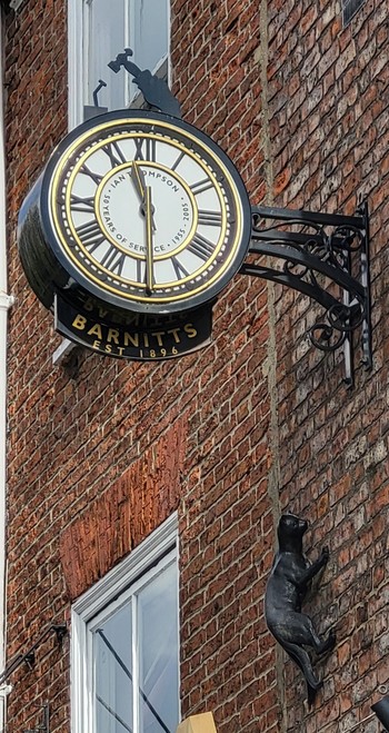 Colour photo of an old clock fixed to a red brick building with a sculpture of a cat walking up to the clock. 
The time says 11.30. At 1, the cat strikes for the mouse!