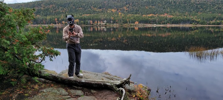 A human dog trying to take a picture with his phone in front of a pond that reflects sky and colorful trees in a green hill
