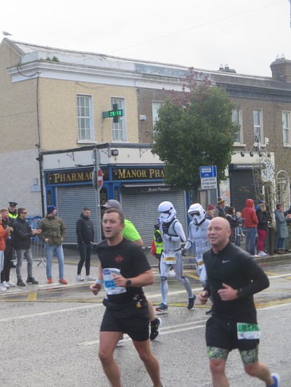 Athletes running along a city street. In the background are two people in costume, one as an Imperial Stormtrooper from the Star Wars films and other as one of the forest scouts from Return of the Jedi.