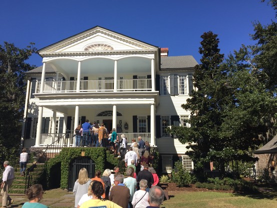 Color image of participants in an annual house tour in Coastal Carolina, USA, queued up to visit. Image shows a graceful antebellum home's two welcoming porches and typical mid-19th century plantation home architecture. The sky is a cloudless blue, and a graceful set of brick stairs cascade down from the lower of the two stacked verandas.