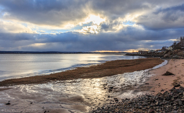 A sandy and muddy shoreline starts lower left of the frame and curves around towards upper right then towards the middle again. The mud bottom left of the frame is shiny and wet, while the surface of the water is only slightly rippling, barely discernible. There are a few older houses in the distance, up on a higher level, middle right of the fram. The sky above the distant shoreline, across the bay has a streak of sunset, below dark clouds that are broken by some very bright light patches near the top of the photo.