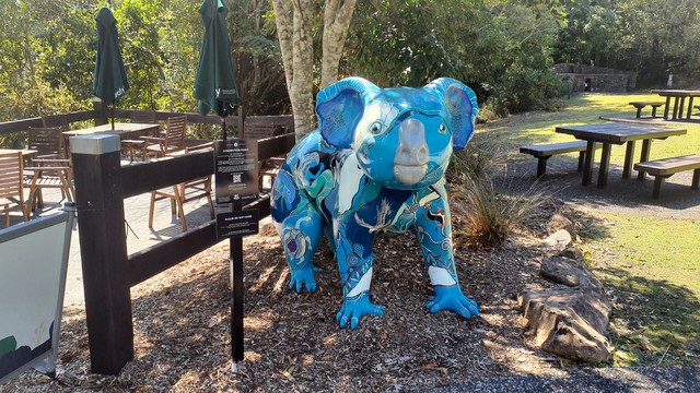 A statue of a koala (with Aboriginal-style artwork, blue in colour), about 1.5m high, stands on all fours beside a sign about wildlife conservation.