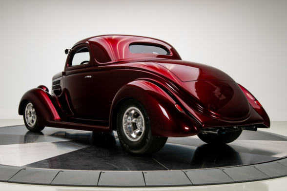 a photo of a 1936 Ford Coupe, seen from behind in a three quarter view, it is a deep burgundy color, the curves of it are very smooth and graceful
