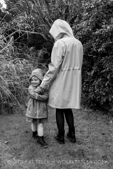 This upright black & white photograph was created in my garden. It shows a young woman and her child daughter, both wearing raincoats. Mother is on the right, daughter on the left. The mother has her back turned towards the camera and is looking away. Her daughter faces the camera but is looking to the side. She is smiling and holds on tightly to her mother’s hand.