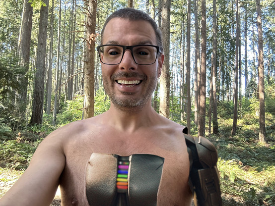 a sexy smiling android is in the woods shirtless. its cheat panel is open revealing metal battery coverings and a status light bar showing a rainbow of colors. also, the skin of its arm has been removed. the droid has a dorky vacant smile and glasses on