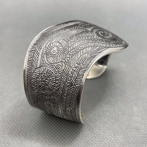 A wide silver bracelet cuff with a curved outline. The surface has been etched with a complex pattern of thin lines and spirals and parallel lines.