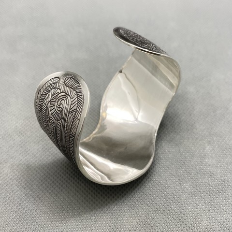 A wide silver bracelet cuff with a curved outline. The surface has been etched with a complex pattern of thin lines and spirals and parallel lines. This is the view of the inside.