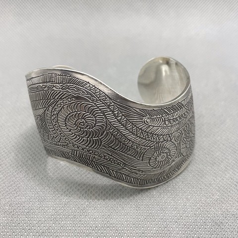 A wide silver bracelet cuff with a curved outline. The surface has been etched with a complex pattern of thin lines and spirals and parallel lines.
