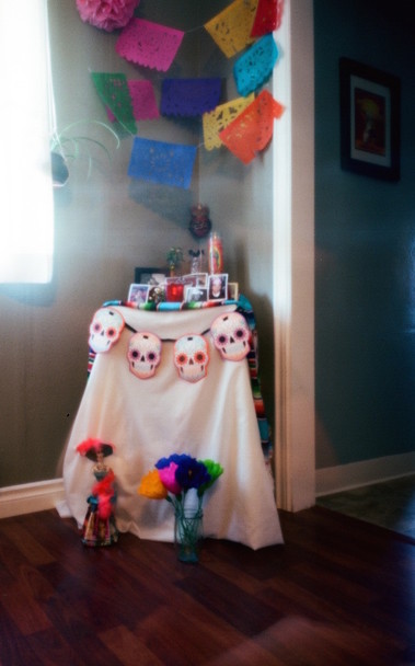 Colorful pinhole photo of the altar in the corner of a room with colorful flags above a small table with photos, candles and paper skulls below and soft light coming in from the left.