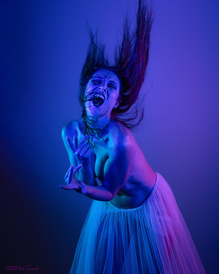 Model Astrid Kallsen poses mid-hair-flip, with her face contorted in a scream. She is cast in blue and purple light. She is topless and wearing a sheer dress wrapped around her waist.