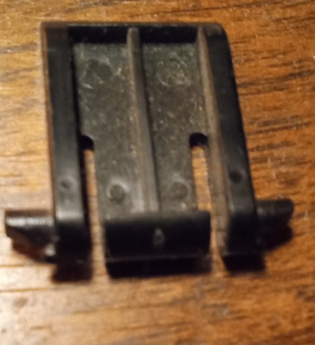 A flat rectangle of plastic with three lateral ribs, two slots are cut halfway the length between ribs, and a cylindrical pin on each side of the bottom end. Black, laying on wood