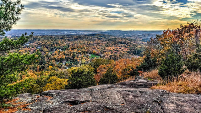 Atop cracked gray stone cliffs under a mostly cloudy sky with thin hazy clouds and patches of deep blue. To the left is a pine tree, To the right and below is a forest of mostly hardwoods and  sprinkling of cedars. A mostly wooded valley hundreds of feet below with glimpses of many houses and other buildings rolls downward towards the city of New Haven on the horizon with Long Island Sound and Long Island just visible as streaks of gray beyond. The forests along our cliff and the peak across the valley sport foliage of oranges and golds, with a scattering of deep green evergreens for contrast.