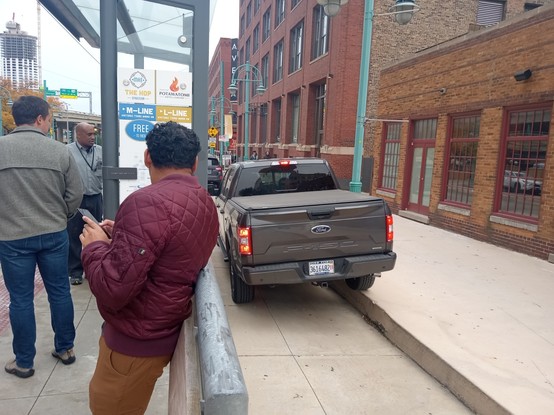 Pickup truck driving down a protected bike lane between a streetcar stop and the sidewalk. The right half of the truck has climbed onto the sidewalk.