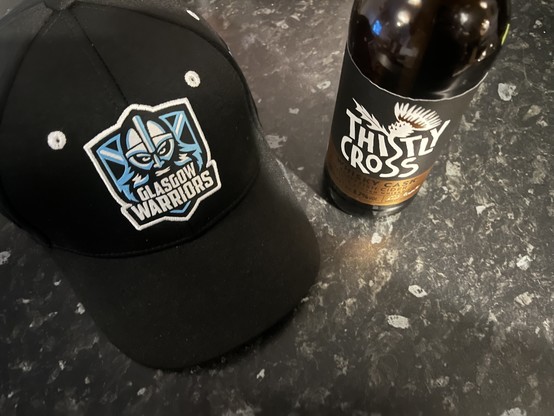 A Glasgow Warriors baseball cap sitting on a counter next to a bottle of Thistly Cross whisky cask aged cider.