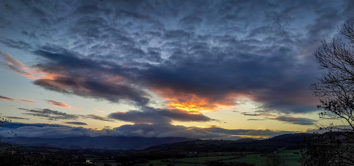 Wide angle shot of dawn/sunrise over Cumbrian hills with farmland in foreground and spectacular clouds above with orange glow of sun shining through