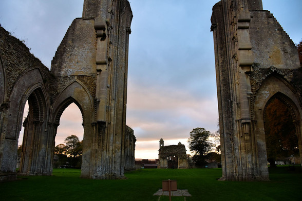 Looking between  two towering stone walls with arches at their base, the remains of a large Medieval church, towards a small chapel building, with  the sky behind in shades of blue and pink and glowing bright near the horizon as the sun sinks