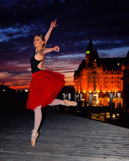 A model in a black body suit, red skirt, dance tights, and point shoes strikes a pose on a wooden boardwalk. In the background is the end of a sunset and beginning of blue hour, with a few clouds, and Ottawa's Chateau Laurier, lit up.