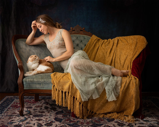 Model Lucy Artmodel reclines on a settee, petting Gus, a cameo tabby Scottish Fold cat. Lucy is wearing a sheer teal vintage gown. Lucy and Gus are making eye contact with each other.