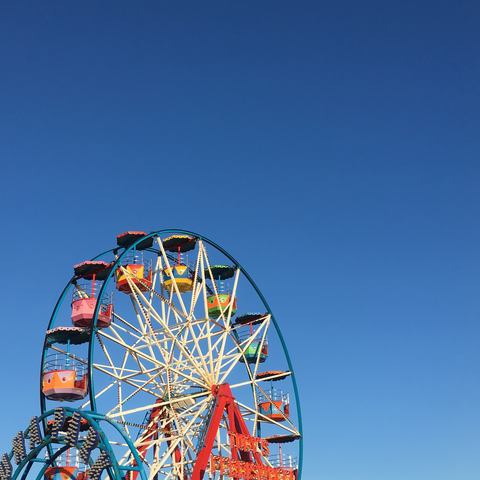 Photo of a Ferris wheel set against a bright blue clear sky. The wheel is in the bottom left hand corner of the image. The spokes of the wheel are white and it has multicoloured cars or seats. The wheel is supported on a red steel base with an illuminated sign on the front which says ‘Tuby’s Ferris Wheel’.