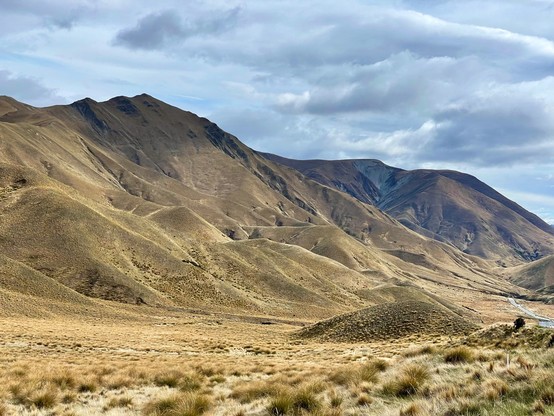 Tussock grass covers the ranges of Lindis Pass; shadows from passing clouds darken the landscape selectively; state highway is visible right of frame
