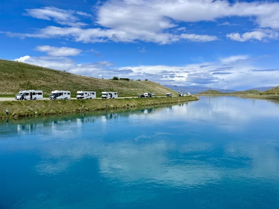 A blue sky reflects in the sediment-rich alpine waters of a power station outflow canal; left of frame a line of vans takes advantage of free overnight parking, while some campers try their luck fishing for salmon