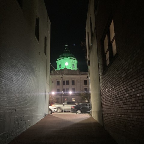 Seen through an alley at night, a courthouse dome is lit with green light.