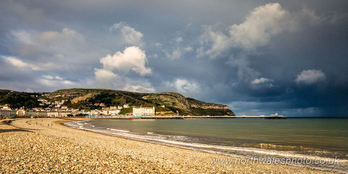 A wide view of a stony beach with a pier and hotel backed by a headland, all in sunshine. Ominous black clouds in the background.