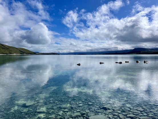 Clouds and blue sky reflected in clear shallow water of lake; stones of the lakebed are readily seen; a row of stones is above water a little way into the water