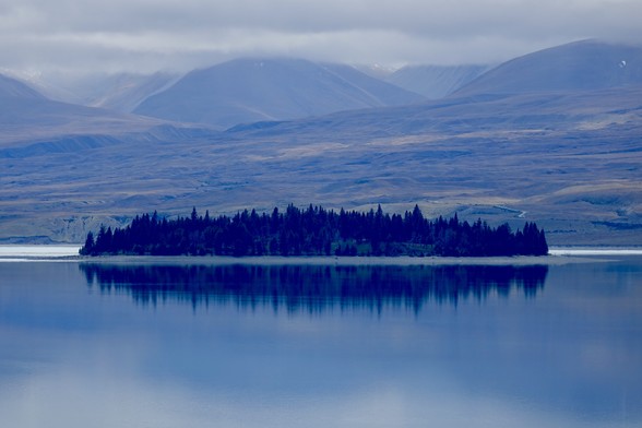 Motuariki Island, looking dark due to lighting conditions, is reflected in the lake; ranges beyond